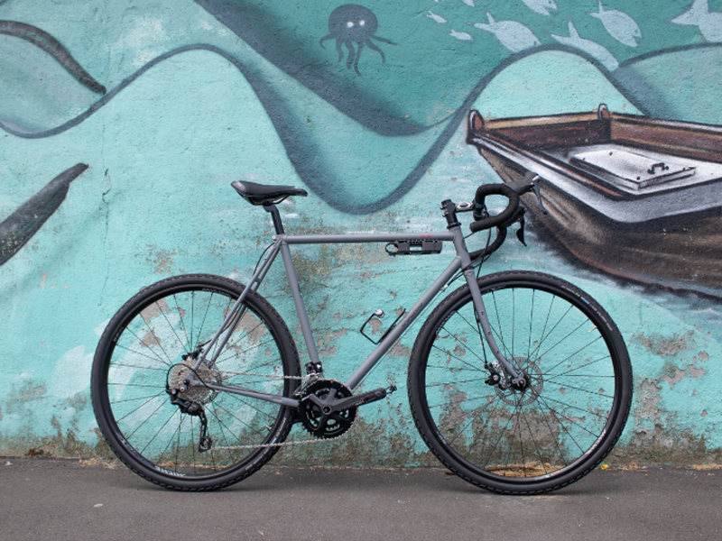 la Gravel Serie X grigia in acciaio di Boeris Bikes Torino fotografata al parco Dora appoggiata a un murales azzurro