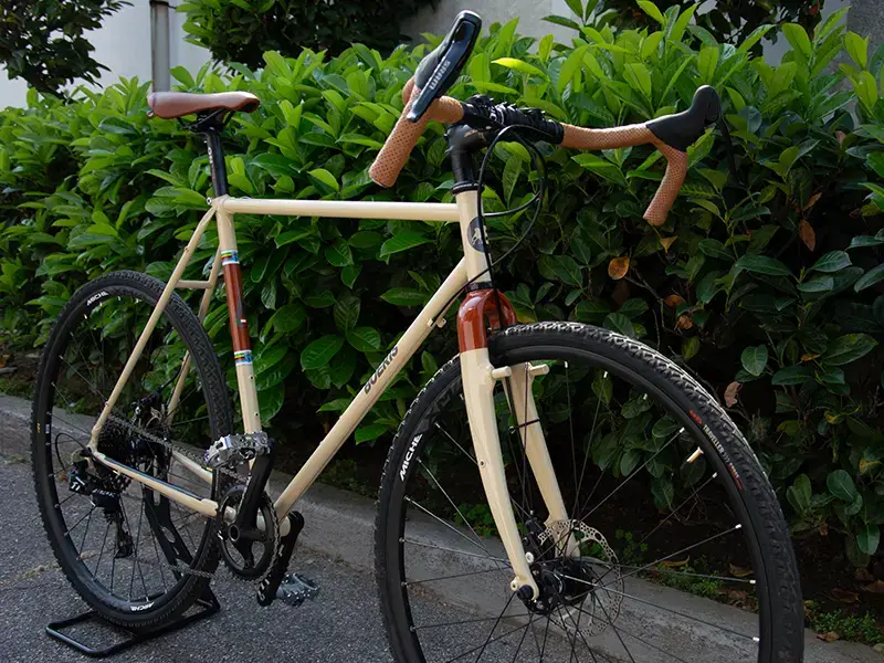 Gravel bike color panna con sella e manubrio marroni di Boeris Bikes Torino di fronte ad una siepe